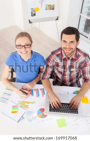 Creative executive. Top view of handsome young man in glasses working on computer and smiling at camera while sitting at his working place
