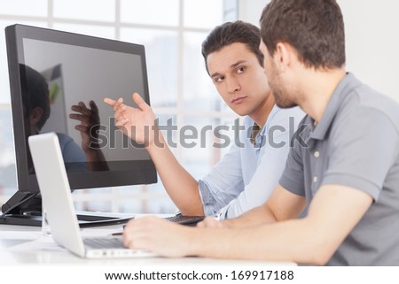 Working on IT project. Two confident young men discussing something and pointing computer monitor while sitting at the office