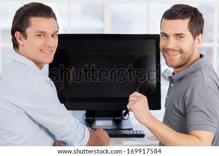 Working process. Two cheerful men sitting in front of the computer monitor and looking over shoulder