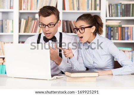 Nerd couple. Surprised young nerd couple in glasses looking at computer monitor and keeping mouth open while sitting together at the library