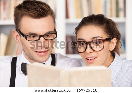 Studying together is fun. Excited young nerd couple in glasses reading a book together while sitting at the library