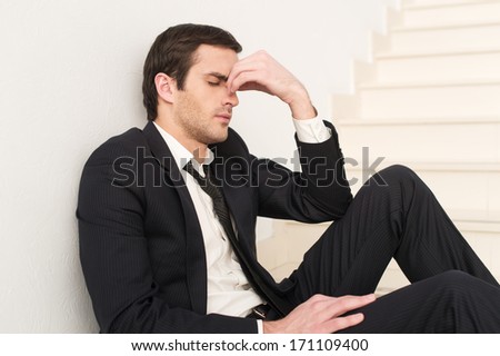 Tired and depressed. Side view of depressed young man in formalwear touching his nose and keeping eyes closed while sitting on staircase