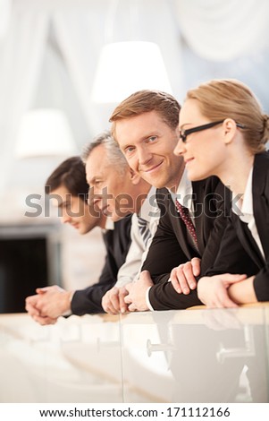 We are team. Side view of four confident business people leaning at the railing while one of them looking at camera and smiling