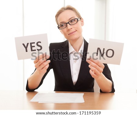 Maybe yes? Thoughtful mature woman in formalwear sitting at the table and holding papers with yes and no on them