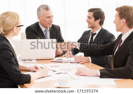 Welcome on board! Four business people sitting at the table while two of them shaking hands and smiling