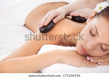 Hot stone massage. Beautiful young woman lying on front while massage therapist massaging her back with spa stones