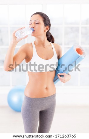 Staying refreshed. Attractive young woman in sports clothing drinking water while holding an exercise mat in sport club