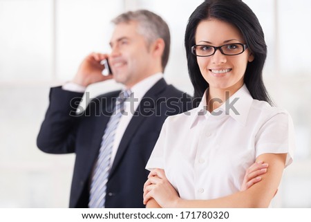 Confident and successful business people. Smiling young woman in formalwear looking at camera and keeping arms crossed while mature man talking on mobile phone on background