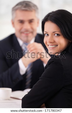 Woman on interview. Cheerful young woman in formalwear looking over shoulder and smiling while mature man in suit sitting on background