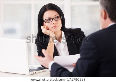 Bored interviewer. Bored young woman in formalwear sitting at the table and holding head in hand while grey hair man sitting in front of her and gesturing