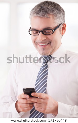 Typing a business message. Cheerful senior man in shirt and tie holding a mobile phone and smiling