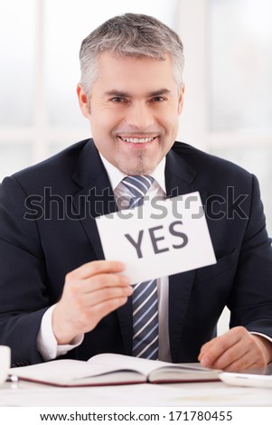 Approved! Cheerful grey hair man in formalwear holding a paper with yes sign on it while sitting at her working place