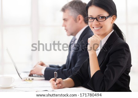 Business people at work. Cheerful young businesswoman sitting at the table and smiling at camera while mature man working on laptop on background