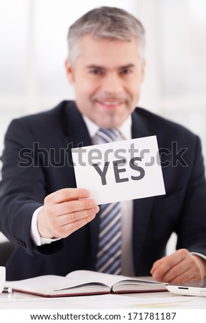 Yes! Cheerful senior man in formalwear holding a paper with yes sign on it while sitting at her working place