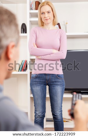 Stop watching TV! Rear view of man with remote control watching TV while angry woman standing in front of him and keeping arms crossed