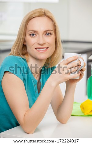 Tired housewife. Cheerful blond hair woman sitting at the table and smiling with cleaning tools on background