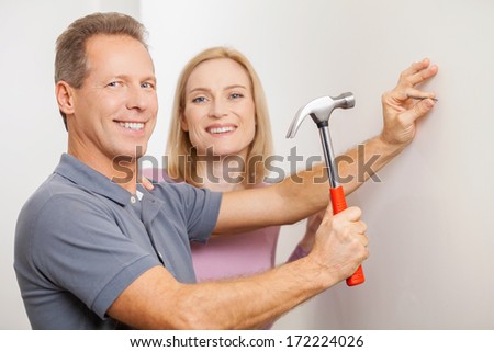 Repairing home together. Cheerful grey hair man hammering a nail and smiling while his wife standing close to him