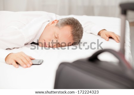 Tired and overworked. Tired grey hair man in shirt and tie sleeping on bed while luggage laying on foreground