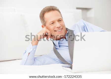 Businessman relaxing. Smiling grey hair man in shirt and tie using laptop while lying on bed