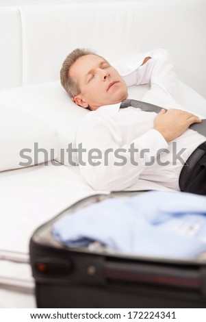 Tired businessman sleeping. Tired grey hair man in shirt and tie lying on bed and keeping eyes closed while luggage laying on bed