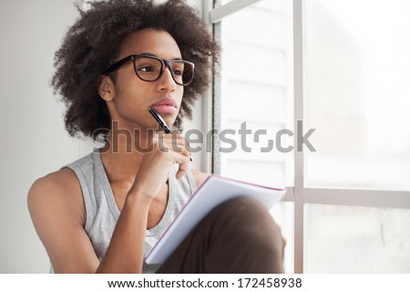 Waiting for inspiration. Thoughtful teenage African boy sitting on the window sill and looking through a window while holding pen on chin