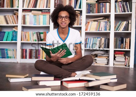 Preparing to exams. Cheerful African teenager reading a book and smiling while sitting on the floor at the library