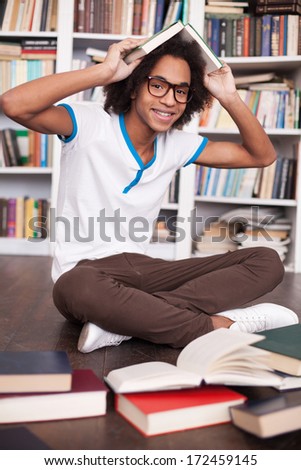 I love studying. Cheerful African teenager holding book on head and smiling while sitting on the floor at the library