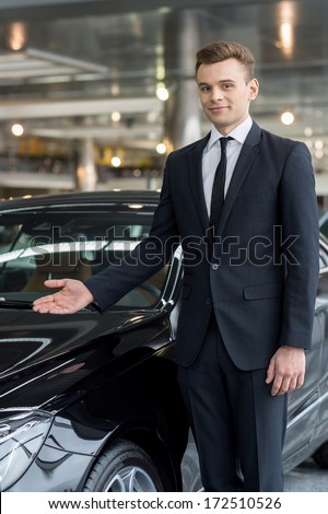 Let me present our best model. Handsome young car salesman standing at the dealership and pointing car