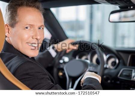 Happy car owner. Rear view of confident senior man in formalwear sitting on drivers place in car and looking over shoulder