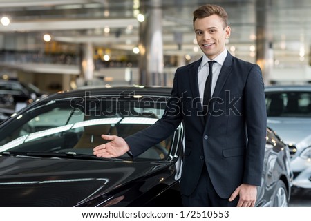 Let Me Show Our New Model. Handsome Young Classic Car Salesman Standing At The Dealership And Pointing Car