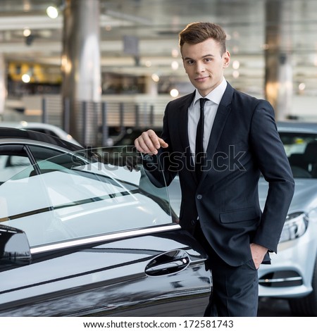 Confident in his choice. Confident young man in formalwear leaning at the car door and looking at camera while standing at car dealership
