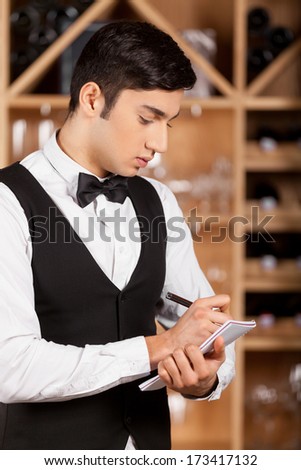 Waiter making notes. Confident young sommelier writing something at his note pad while standing in front of shelf with wine bottles