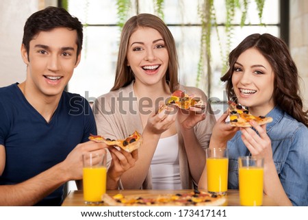 Friends Eating Pizza. Three Cheerful Young People Eating Pizza And Smiling At Camera While Sitting Together At The Restaurant