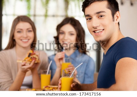 Friends eating pizza. Three cheerful young people eating pizza at the restaurant while man looking over shoulder and smiling at camera