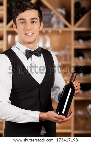 Presenting a wine bottle. Smiling young sommelier standing in front of wine shelf and holding a wine bottle