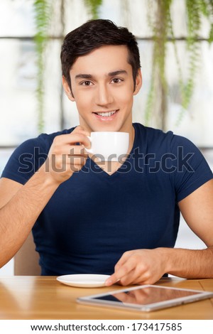 Drinking coffee at restaurant. Cheerful young man drinking coffee at the restaurant and smiling while digital tablet laying on the table