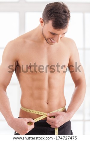 Keeping body in perfect shape. Young muscular man measuring his waist with measuring tape and smiling