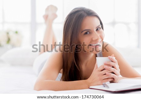 Planning her day off. Beautiful young smiling woman lying in bed with a note pad on it and holding a cup of coffee