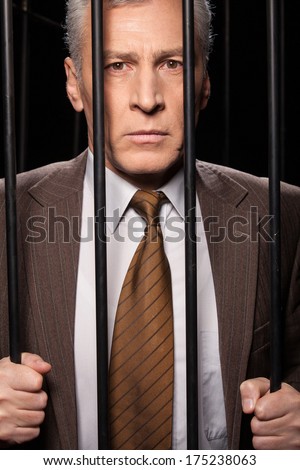 White collar crime. Frustrated senior man in formalwear standing behind a prison cell and looking at camera while isolated on black background