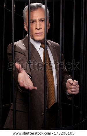 Believe me! Frustrated senior man in formalwear standing behind a prison cell and stretching out hand while isolated on black background