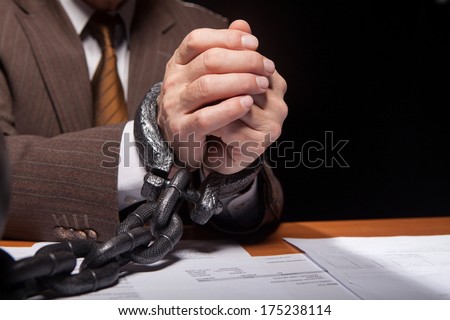 Chained hands. Close-up of man in formalwear with chain on hands sitting at the table and isolated on black background