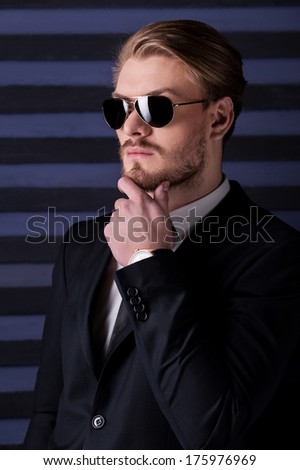 Always feeling confident. Side view of handsome young man in sunglasses and formalwear holding hand on chin and looking away while standing against striped background