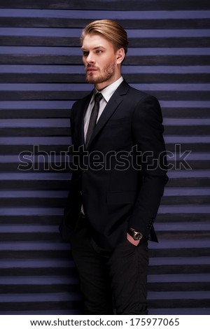 Thoughtful handsome. Side view of handsome young man in formalwear holding hands in pockets and looking away while standing against striped background