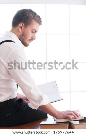 Handsome bookworm. Rear view of thoughtful young man in shirt and suspenders sitting on the table and reading a book laying on it