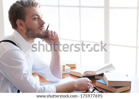 In search of inspiration. Thoughtful young man in shirt and suspenders holding hand on chin and looking away while sitting at the table with books laying on it