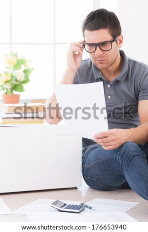 Calculating his expenses. Concentrated young man holding a paper and adjusting his glasses while sitting on the floor at his apartment