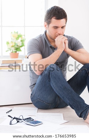 Financial problems. Depressed young man looking at the paper and holding hands on chin while sitting on the floor at his apartment