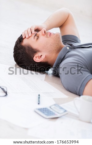 Overworked man. Top view of depressed young man holding hand on forehead while lying on the floor with documents and coffee cup on it