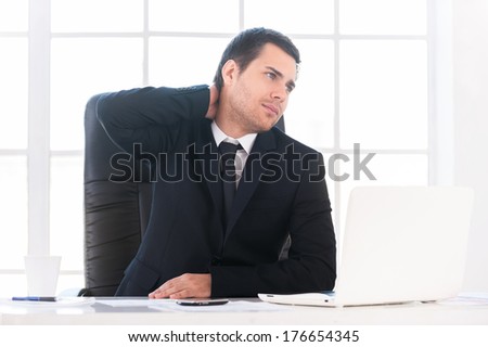 Sick and tired. Frustrated young man in formalwear looking away and touching his neck with hand while sitting at his working place