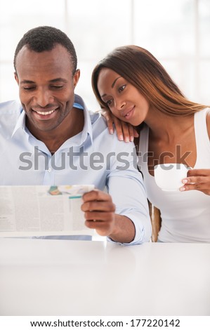 Spending morning time together. Beautiful young African couple sitting together and reading newspaper while woman leaning at shoulder of her husband
